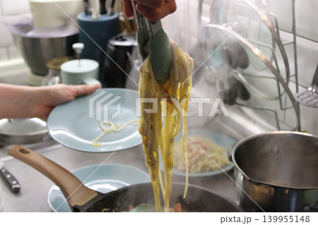 Person holding tongs with cooked spaghetti above a blue plate in a kitchen, steam rising from a pot on the stove, utensils and appliances visible in the background Person holding tongs with cooked spaghetti above a blue plate in a kitchen, steam rising from a pot on the stove, utensils and appliances visible in the background 139955148
