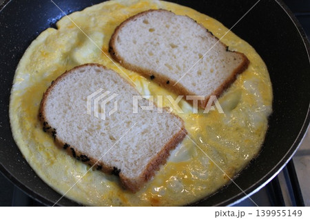 Slices of bread placed on top of a fluffy omelette cooking in a non-stick frying pan on a stovetop, with visible heat source and kitchen background Slices of bread placed on top of a fluffy omelette cooking in a non-stick frying pan on a stovetop, with visible heat source and kitchen background 139955149