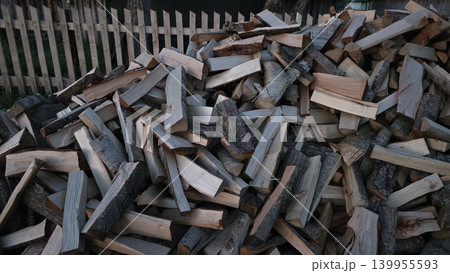 Large pile of chopped firewood lying outdoors near wooden fence in countryside, concept of preparation for winter heating season Large pile of chopped firewood lying outdoors near wooden fence in countryside, concept of preparation for winter heating season 139955593