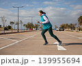 Woman running in full stride performs sport workout session in open air parking lot near sea on sunny day. Outdoor running exercise captures dynamic movement with teal vest and green leggings 139955694