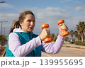 Woman exercising with orange dumbbells performs curl movement during workout session along coastal promenade. Outdoor fitness training captures active movement with arms raised and focused expression 139955695