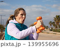 Woman exercising with orange dumbbells shows effort expression during workout along coastal promenade. Outdoor fitness training captures exertion moment with both arms raised and mouth open exhaling. 139955696