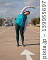 Woman 45 years old stretching sideways raises one arm overhead during sport workout outdoors in parking lot near sea. Outdoor exercise session focuses on lateral flexibility and warm up on sunny day. 139955697