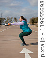 Woman doing squat exercise holds orange dumbbells extended forward during sport workout outdoors in parking lot. Side view of workout taking place near sea on sunny day in open air setting. 139955698