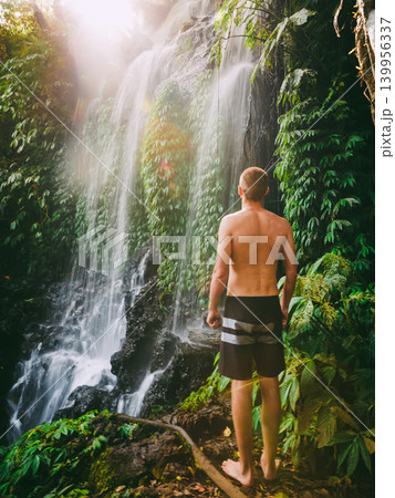 Male traveler standing beside tropical waterfall in lush jungle landscape 139956337