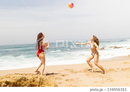 Two women playing with a colorful beach ball on sandy shore near ocean waves, wearing swimsuits under bright sunlight with rocky formations in the background 139956381