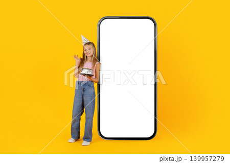 Smiling little girl standing beside oversized smartphone mockup with birthday cake Smiling little girl standing beside oversized smartphone mockup with birthday cake 139957279