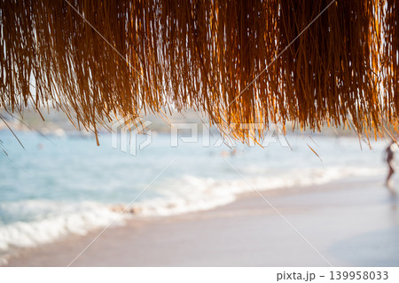 Straw beach umbrella hangs above sandy shoreline with blurred sea background during sunny summer vacation. Beach resort lifestyle tropical relaxation and seaside tourism Straw beach umbrella hangs above sandy shoreline with blurred sea background during sunny summer vacation. Beach resort lifestyle tropical relaxation and seaside tourism 139958033
