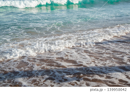 Sea waves wash over sandy beach with white foam and turquoise water during sunny summer vacation weather. Beach resort lifestyle coastal relaxation and seaside tourism 139958042