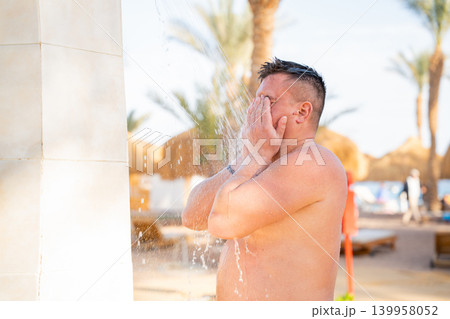 Caucasian man washes face under outdoor beach shower near tropical resort during sunny summer vacation weather. Beach resort lifestyle hygiene refreshment and seaside tourism Caucasian man washes face under outdoor beach shower near tropical resort during sunny summer vacation weather. Beach resort lifestyle hygiene refreshment and seaside tourism 139958052