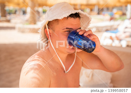 Caucasian man drinks water from blue cup while resting on a beach in Sharm El Sheikh Egypt during summer vacation. Beach resort lifestyle hydration and leisure tourism on the Red Sea 139958057