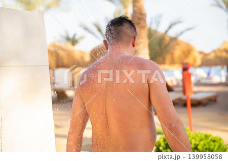 Caucasian man rinses under outdoor beach shower near tropical resort during sunny summer vacation weather. Beach resort lifestyle hygiene relaxation and seaside tourism 139958058