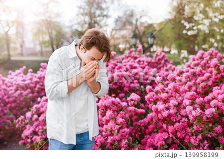 Man outdoors sneezing into a tissue due to spring allergy season and hayfever. He reacts to pollen from the vibrant pink blooms and flowers. 139958199