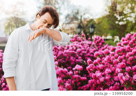 Sick man outdoors suffering from spring allergy and hayfever. He is sneezing into his elbow due to pollen from the bright pink blooms and flowers. Sick man outdoors suffering from spring allergy and hayfever. He is sneezing into his elbow due to pollen from the bright pink blooms and flowers. 139958200