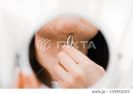Reflection in a mirror of unwanted facial hair removal from the chin using a tool. Woman doing a home beauty routine for hair growth due to hormonal changes. 139958203