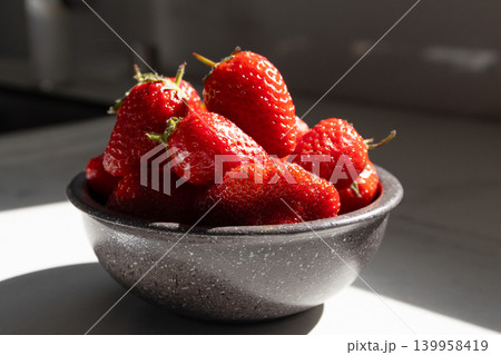 Close-up of ripe strawberries in gray bowl under soft daylight. Fresh red berries with green leaves on white surface. Healthy fruit concept for summer snacks and organic eating. Close-up of ripe strawberries in gray bowl under soft daylight. Fresh red berries with green leaves on white surface. Healthy fruit concept for summer snacks and organic eating. 139958419