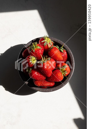 Fresh strawberries in black bowl on white surface with sunlight shadow. Top view flat lay composition highlighting juicy red fruit texture and natural Fresh strawberries in black bowl on white surface with sunlight shadow. Top view flat lay composition highlighting juicy red fruit texture and natural 139958420
