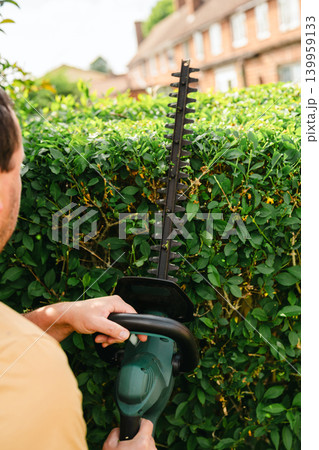 Person trimming garden hedge with electric hedge trimmer, backyard gardening maintenance Person trimming garden hedge with electric hedge trimmer, backyard gardening maintenance 139959133