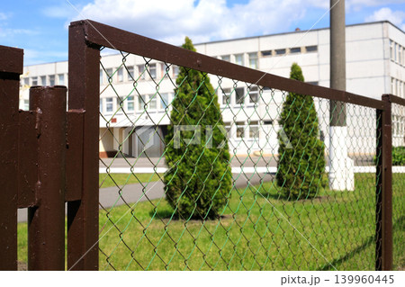 Metal chain link fence with brown steel posts on green lawn background, wire mesh boundary grid with thuja trees near school building Metal chain link fence with brown steel posts on green lawn background, wire mesh boundary grid with thuja trees near school building 139960445