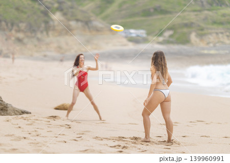 Two women play frisbee on a sandy beach during a sunny day while waves crash in the background and people relax around them Two women play frisbee on a sandy beach during a sunny day while waves crash in the background and people relax around them 139960991