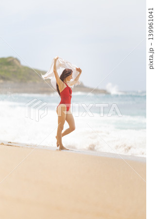 Woman enjoys the beach and waves while holding a scarf on a sunny day in summer near the ocean shoreline Woman enjoys the beach and waves while holding a scarf on a sunny day in summer near the ocean shoreline 139961041