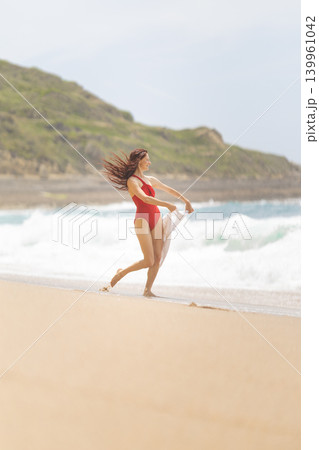 Woman walks on sandy beach near ocean waves while holding fabric in the air during sunny day at coastal area Woman walks on sandy beach near ocean waves while holding fabric in the air during sunny day at coastal area 139961042