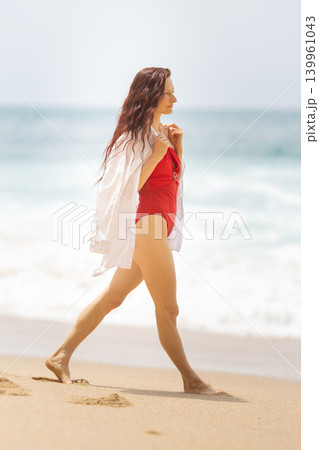 Woman walks along the beach in swimwear with a cover-up during a sunny day by the ocean 139961043