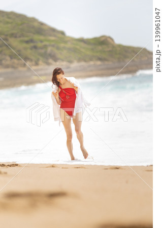 Woman in red swimsuit walking along the sandy beach at the shore on a bright sunny day near the waves Woman in red swimsuit walking along the sandy beach at the shore on a bright sunny day near the waves 139961047