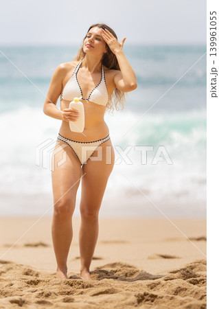 Woman walks on beach while holding sunscreen and enjoying the sun at the ocean in daylight Woman walks on beach while holding sunscreen and enjoying the sun at the ocean in daylight 139961055