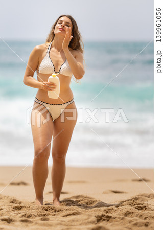 Woman applying sunscreen on a beach while walking on the sand near the ocean in the afternoon sun 139961056