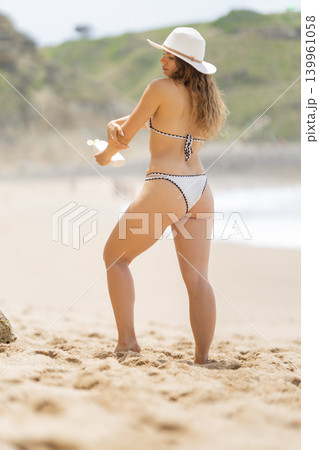 Woman applies sunscreen on the beach during summer with a sun hat while standing in soft sand near the ocean 139961058