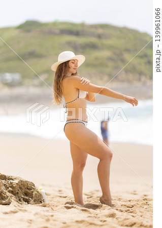 Woman in a bikini enjoys a sunny day at the beach while applying sunscreen near the ocean shore with other beachgoers around Woman in a bikini enjoys a sunny day at the beach while applying sunscreen near the ocean shore with other beachgoers around 139961066
