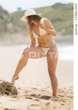 Woman applies sunscreen while standing on the beach on a sunny day near the ocean Woman applies sunscreen while standing on the beach on a sunny day near the ocean 139961068