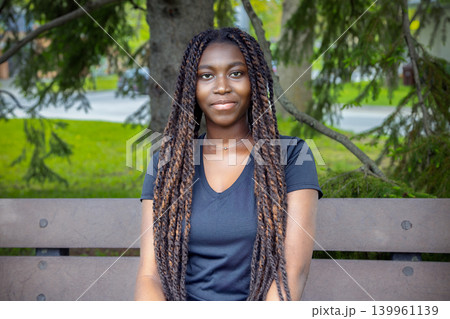 Young African woman sitting on park bench, summer outdoor portrait 139961139