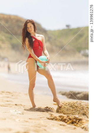 Woman stands on sandy beach holding a blue ball with waves in background during sunny day near rocky shore 139961276