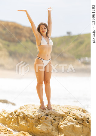 Female model in white bikini poses on a rock at the beach, arms raised, with ocean waves and green hills in the background under a bright sky Female model in white bikini poses on a rock at the beach, arms raised, with ocean waves and green hills in the background under a bright sky 139961292