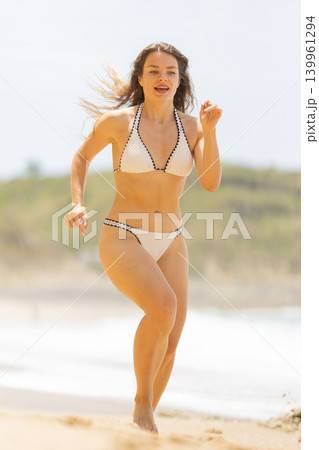 Female model in white bikini running along sandy beach with ocean waves and green hills in the background under a bright sunny sky Female model in white bikini running along sandy beach with ocean waves and green hills in the background under a bright sunny sky 139961294