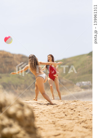 Two women playing with a beach ball on sandy shore, one in red swimsuit and the other in white bikini, with green hills and blue sky in the background Two women playing with a beach ball on sandy shore, one in red swimsuit and the other in white bikini, with green hills and blue sky in the background 139961311