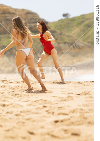 Two young women running on sandy beach, one in red swimsuit and the other in white bikini, with green hills and ocean waves in the background under bright sunlight Two young women running on sandy beach, one in red swimsuit and the other in white bikini, with green hills and ocean waves in the background under bright sunlight 139961316