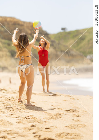 Two women playing with a beach ball on sandy shore, one in a red swimsuit and the other in a bikini, with ocean waves and green hills in the background Two women playing with a beach ball on sandy shore, one in a red swimsuit and the other in a bikini, with ocean waves and green hills in the background 139961318
