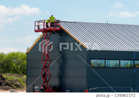 Workers on scissor lift installing metal roof panels on modern industrial building construction Workers on scissor lift installing metal roof panels on modern industrial building construction 139961572