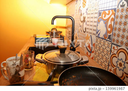 Messy kitchen counter with dirty dishes, pots, and pans after cooking. Domestic chaos in a modern kitchen with patterned tiles and a black faucet Messy kitchen counter with dirty dishes, pots, and pans after cooking. Domestic chaos in a modern kitchen with patterned tiles and a black faucet 139961987