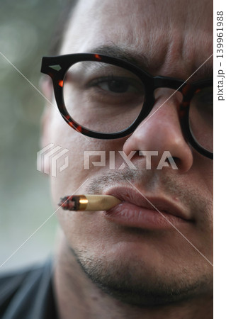 Close up portrait of a young man in glasses smoking a cigarette. Cinematic male face with serious expression and blurred background Close up portrait of a young man in glasses smoking a cigarette. Cinematic male face with serious expression and blurred background 139961988