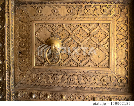 The fragment depicts a gilded wooden door decorated with fine carvings and a bronze ring. Traditional Indian decorative art, Jaipur. The fragment depicts a gilded wooden door decorated with fine carvings and a bronze ring. Traditional Indian decorative art, Jaipur. 139962183