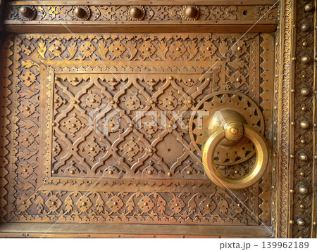 The fragment depicts a gilded wooden door decorated with fine carvings and a bronze ring. Traditional Indian decorative art, Jaipur. 139962189
