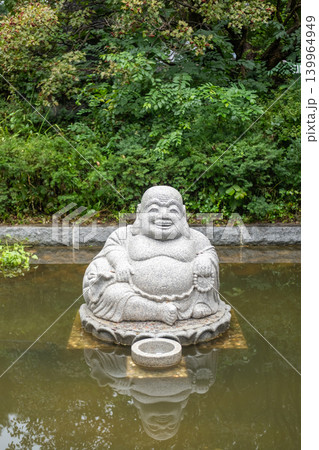 Stone Budai, or Laughing Buddha statue sitting in a shallow pond at Bongeunsa Temple in Seoul, surrounded by calm water and lush greenery. 139964949