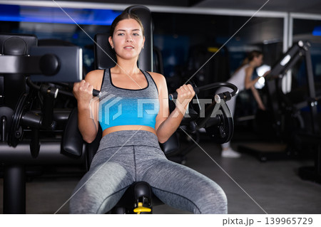 Sportive young girl training on arm curl machine in gym Sportive young girl training on arm curl machine in gym 139965729