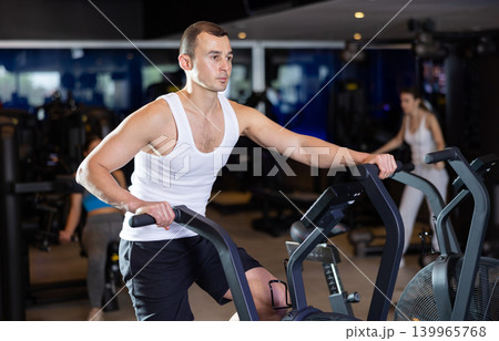 Sportive young guy doing air bike exercises in well-equipped gym Sportive young guy doing air bike exercises in well-equipped gym 139965768