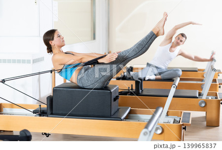 Girl works out on in gym on Pilates simulator. Sports club client on reformer with cable and rope performs exercise to stretch muscles of arms in gym Girl works out on in gym on Pilates simulator. Sports club client on reformer with cable and rope performs exercise to stretch muscles of arms in gym 139965837