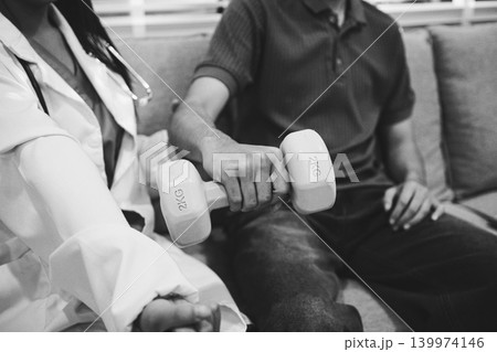 An Asian male doctor and his patient are seated in chairs, performing physical therapy using dumbbells under the doctor's supervision. The patient is experiencing pain. An Asian male doctor and his patient are seated in chairs, performing physical therapy using dumbbells under the doctor's supervision. The patient is experiencing pain. 139974146